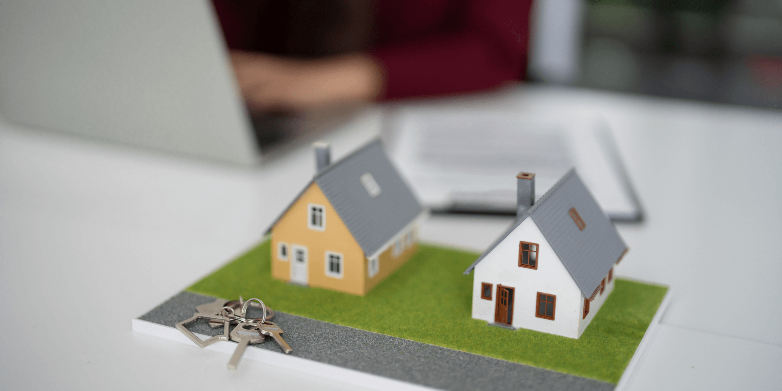 Model houses on a desk with keys in front and a person working on a laptop in the background.